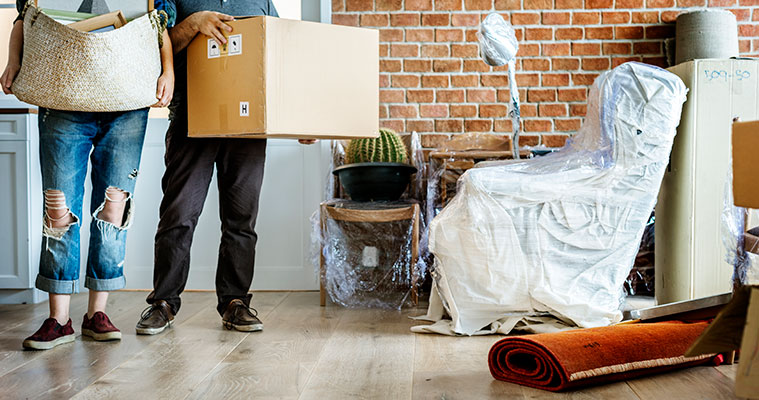 Dos personas en una habitación transportando cajas y cestas con objetos personales, rodeados de muebles envueltos, plantas, una alfombra enrollada y una lámpara, ilustrando el proceso de guardar muebles