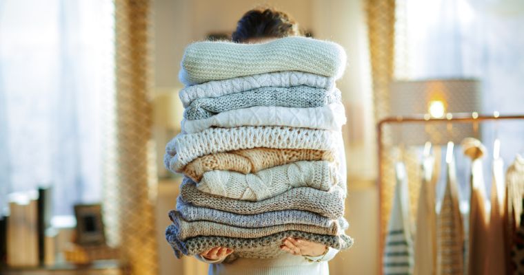 Persona cargando una pila de jerséis con un perchero al fondo, ilustrando consejos para guardar ropa durante mucho tiempo sin perder calidad
