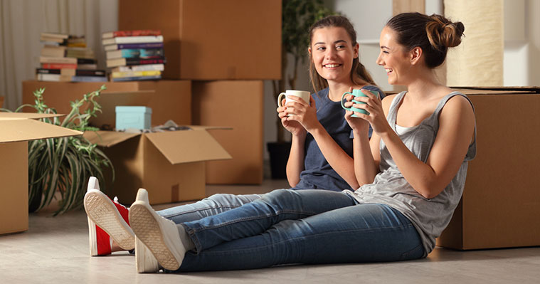Dos jóvenes sentados en el suelo con tazas de café, rodeados de cajas, libros y plantas, representando una mudanza estudiantil