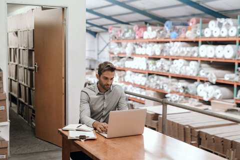 Business owner working on a laptop inside a storage unit with flooring materials and carpet rolls, showing flexible OhMyBox business storage.
